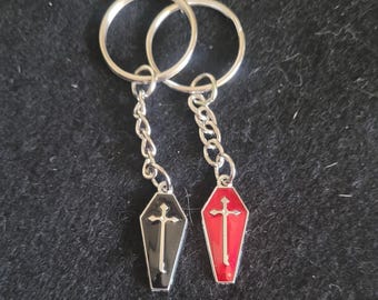 May include: Two keychains with coffin-shaped pendants. One pendant is black with a silver cross, the other is red with a silver cross. Both pendants are attached to silver chains and key rings.