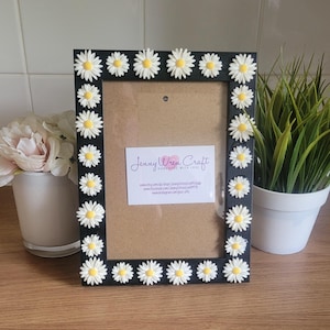 May include: Black picture frame with a white daisy border. The frame has a blank space for a photo and a small card with the text "Jenny Wren Craft Handmade with Love" and website information.