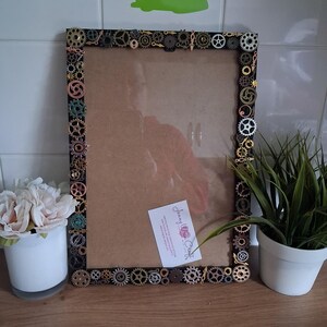 May include: A rectangular picture frame with a steampunk design. The frame is black and decorated with various metal gears in shades of gold, bronze, and silver. The frame holds a brown backing and a small white card with cursive text.