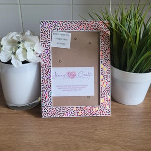 May include: A white picture frame with a pink and red glitter border. The frame has a brown cardboard backing with the text "Jenny Wren Craft Handmade with Love" and contact information. The text "Remove protective covering from both sides" is visible on a small white sticker.