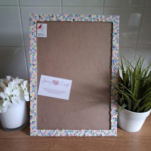 May include: A rectangular picture frame with a colorful, textured border. The border is made of small, round, multi-colored embellishments. The frame has a brown backing and a white label with text. A small plant in a white pot is visible.