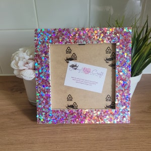 May include: A white picture frame with a pink and iridescent glitter border. The frame is decorated with small star-shaped glitter. The frame is sitting on a wooden surface. The text "Honey Wren Craft" is visible on a business card inside the frame.