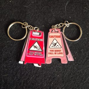May include: Two keychains shaped like wet floor signs. One is bright red and the other is light pink. Both feature a triangular icon of a person slipping with hearts and the text CAUTION YOU MIGHT FALL IN LOVE printed in black and white.