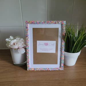 May include: A white picture frame with a colorful confetti border. The frame is 4x6 inches and has a brown cardboard backing. The frame is handmade with love by Jenny Wren Craft.
