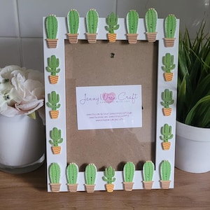 May include: White picture frame with a cactus theme. The frame is decorated with green and yellow cactus illustrations. The frame is 8x10 inches and has a brown mat. The text "Jenny Wren Craft Handmade with Love" is printed on the mat.