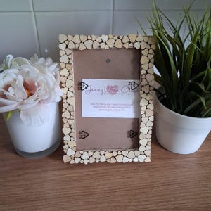 May include: A rectangular picture frame featuring a border covered in small, light-coloured wooden hearts. A white card is centred inside the frame with the text Jenny Wren Crafts, along with social media links and recycling symbols.