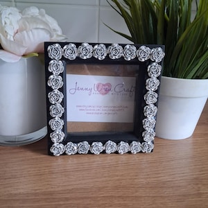 May include: A black square picture frame with silver rose embellishments around the border. The frame has a clear glass insert and a printed card inside with the text "Jenny Wren Craft". The frame is displayed on a wooden surface.