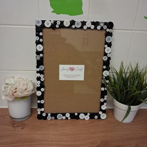 May include: A rectangular black picture frame decorated with an assortment of white and black buttons. The frame has a brown backing and a clear glass front. A small white label with text is affixed to the frame. A small white vase with flowers is on the left.