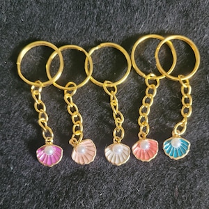 May include: Five gold keyrings with a small seashell charm. The charms are in different colours: pink, light pink, white, coral, and teal.
