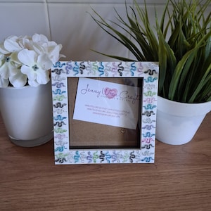 May include: Square white picture frame decorated with colourful snake-like designs. The frame has a clear glass front and a cardboard backing. The frame is sitting on a wooden surface. The frame has a small card inside with the text "Jenny Wren Crafts".