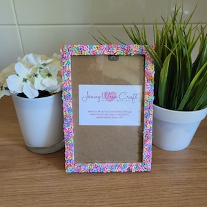 May include: A colorful picture frame with a white background and a pink, blue, yellow, and green sprinkle border. The frame has a hanging hook on the back and a business card inside with the text "Jenny Wren Craft Handmade with Love" and contact information for Etsy, Facebook, and Instagram.
