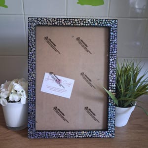 May include: A rectangular picture frame with a black border covered in small, holographic silver stars. The back of the frame is made of brown cardboard with four clear plastic reminder stickers and a white business card attached to the center.