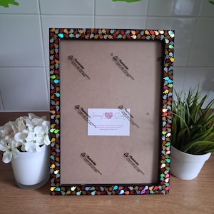 May include: A rectangular black picture frame with a colorful leaf pattern border. The frame has a brown backing and a clear protective sheet. A small white label with text is visible on the frame.