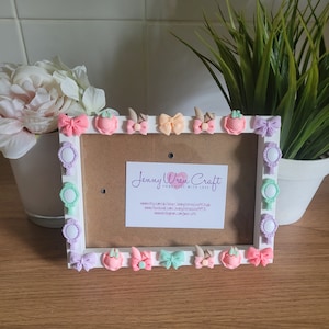 Puede incluir: Marco de fotos blanco decorado con lazos, espejos y otros adornos en rosa pastel, verde y morado. El marco tiene un fondo blanco con el texto "Jenny Wren Craft Handmade with Love" e información del sitio web.