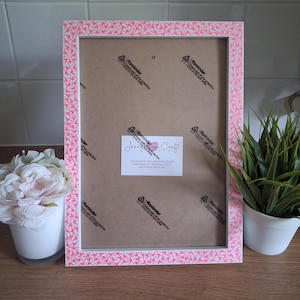 May include: A rectangular picture frame with a white border and a pink pattern of small shapes. The frame has a white interior and a brown backing. A white label with text is visible on the backing. A white vase with flowers is on the left.