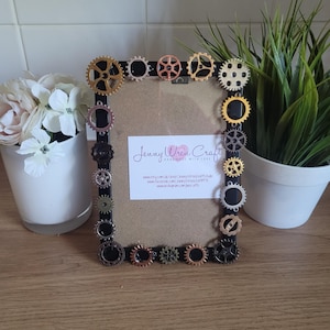 May include: A black picture frame decorated with gold, silver, and copper gears. The frame is surrounded by a white background. The frame has a business card inside with the text "Jenny Wren Craft Handmade with Love" and contact information.
