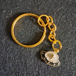 May include: Gold-colored keychain with a small charm shaped like a UFO. The UFO charm has a white top, black trim, and small silver-colored dots. The keychain ring and chain are also gold.