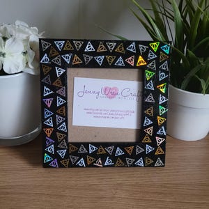 May include: Black picture frame with a gold, silver, and iridescent holographic border featuring a repeating pattern of the Deathly Hallows symbol. The frame has a small white card inside with the text "Jenny Wren Crafts Handmade with Love" and website information.