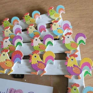 May include: A collection of colourful wooden rooster buttons with rainbow-coloured tails. The buttons are arranged in rows on a white surface.