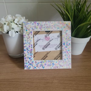 May include: White square picture frame with a white background and a border of iridescent sequins. The frame is sitting on a wooden surface. The frame has a small card inside with the text "Jenny Wren Craft Handmade with Love" and website information.