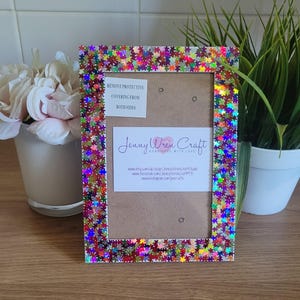 Peut inclure: Un cadre photo blanc avec une bordure colorée, irisée, en forme d'étoile. Le cadre comporte une note indiquant "Retirer le film protecteur des deux côtés". Le cadre comporte également une carte de visite indiquant "Jenny Wren Craft Handmade with Love" et des informations sur le site Web.