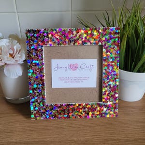 May include: A white picture frame with a colourful iridescent star-shaped glitter border. The frame has a white mount and a card inside with the text "Jenny Wren Craft Handmade with Love" and contact information for the shop.