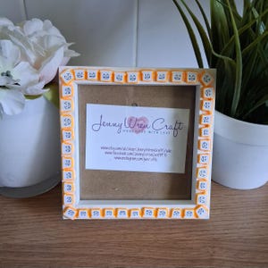 May include: Square white picture frame with orange and white decorative accents. The frame has a small paper insert with the text "Jenny Wren Craft" and social media information.