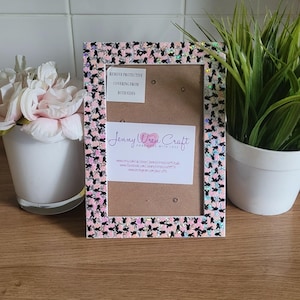May include: A pink and black picture frame with a glittery finish. The frame has a white border and is decorated with a pattern of black bunny silhouettes. The frame has a small white card inside with the text "Jenny Wren Craft Handmade with Love" and contact information.