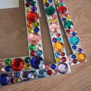 May include: Two rectangular picture frames featuring silver-toned borders densely decorated with an assortment of multi-coloured, faceted acrylic rhinestones in various sizes, including red, blue, green, yellow, orange, pink, and purple gems.