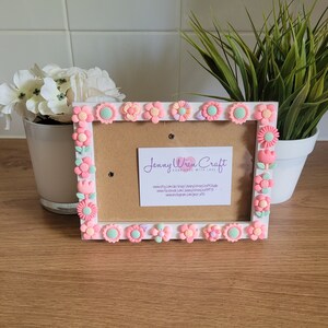 May include: White picture frame decorated with pink, green, and white 3D flower embellishments. The frame has a corkboard backing and a business card inside that reads "Jenny Wren Craft Handmade with Love" with website and social media information.