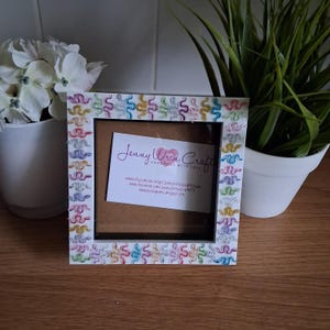 May include: A square white picture frame decorated with colourful squiggly patterns. The frame has a black interior and a small card with the text "Jenny Wren Craft". The frame is displayed on a wooden surface.
