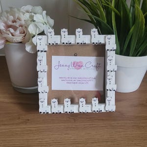 May include: White wooden picture frame decorated with a border of small white llama figurines. The frame has a white mat and a clear glass front. The frame is sitting on a wooden surface. The text on the card inside the frame reads "Jenny Wren Craft Handmade with Love" and includes website addresses for Etsy, Facebook, and Instagram.