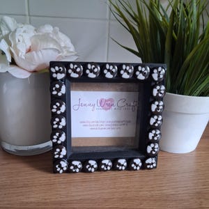May include: A black square photo frame decorated with paw print buttons. The frame contains a small picture with the text "Jenny Wren Craft". The frame is on a wooden surface.