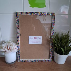 May include: A rectangular picture frame with a border of colorful rhinestones. The frame has a clear glass front and a brown backing. A small white label with text is affixed to the backing. A white pot with pink flowers and a small potted plant are on the surface.