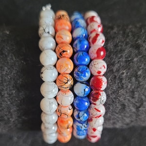 May include: Four beaded bracelets with a variety of colors. The bracelets feature white, orange, blue, and red beads. Each bead has a unique pattern of black lines. The bracelets are stacked together, showcasing the different color combinations.