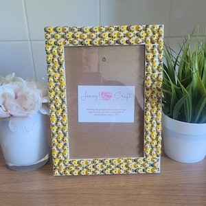 May include: A white picture frame with a yellow and black bee design. The frame is decorated with small, round, 3D bees. The frame is sitting on a wooden surface.