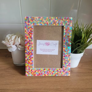 May include: A white picture frame with a colourful star design. The frame is decorated with small, iridescent stars in shades of pink, blue, green, yellow, and orange. The frame is empty and ready to display a photo or artwork. The text "Jenny Wren Craft Handmade with Love" is visible on a card inside the frame.