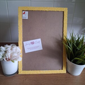 May include: A rectangular yellow picture frame with a decorative border. The frame has a brown backing and a white label with text. A small potted plant and a vase of flowers are on the left and right.
