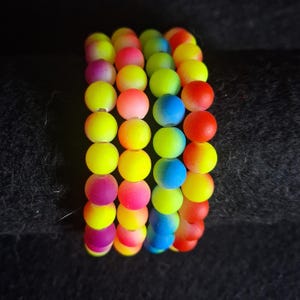 May include: A vibrant bracelet composed of multiple strands of colorful, round beads. The beads transition through a spectrum of colors, including yellow, pink, blue, and red. The bracelet is set against a dark background, highlighting the bright colors.