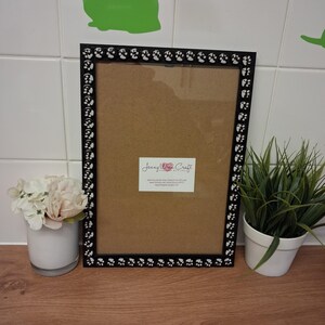 May include: Black picture frame with paw print accents. The frame has a brown backing and a clear glass front. A small white label with text is affixed to the frame. A small white vase with flowers and a potted plant are next to the frame.