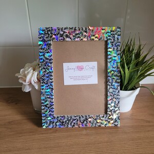May include: A silver picture frame with a black and iridescent glitter border. The frame is sitting on a wooden surface with a small plant in a white pot to the right.