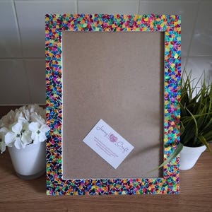 May include: A rectangular picture frame with a colorful sequin border. The frame's border is adorned with a vibrant array of sequins in various colors, including red, yellow, blue, and green. The frame has a neutral-colored backing.