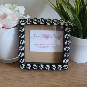 May include: Black picture frame with white paw print embellishments. The frame has a brown mat and a white card inside with the text "Jenny Wren Craft Handmade with Love" and website information.