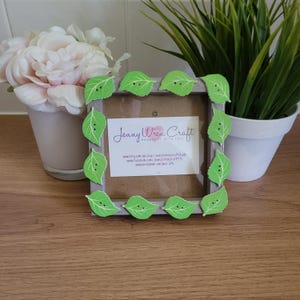 May include: A grey picture frame with a green leaf border. The frame is decorated with green leaves with small white dots. The frame has a card inside with the text "Jenny Wren Craft Handmade with Love" and contact information for the craft business.