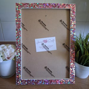 May include: A rectangular picture frame with a glittery, iridescent border. The frame has a white interior and a cardboard backing. A small white label with the text "Jenny Wren Craft" is affixed to the backing.