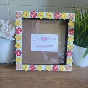 May include: A white square photo frame adorned with yellow and red flower decorations. The frame has a brown backing and a clear glass front. The text "Jenny Wren Craft" is displayed on a white label.