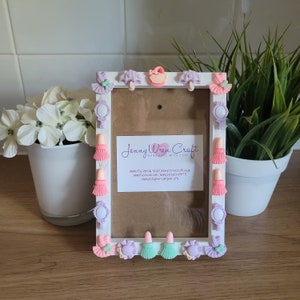 May include: A white picture frame decorated with pastel-coloured embellishments. The frame features miniature lipsticks, mirrors, tutus, and handbags. The frame has a brown backing and includes the text "Jenny Wren Craft Handmade with Love".