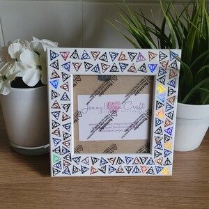 May include: White picture frame with a black and white border featuring a repeating pattern of the Deathly Hallows symbol in various colors. The frame is decorated with a holographic finish. The frame is 4x6 inches.