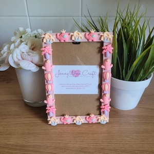 May include: A pink picture frame decorated with 3D embellishments, including elephants, palm trees, hot air balloons, and cacti. The frame has a white background and the text "Jenny Wren Craft Handmade with Love" is printed on the frame.