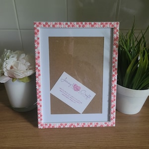May include: White picture frame with a pink and white checkered border. The frame is sitting on a wooden surface. A business card is inside the frame with the text "Jenny Wren Craft Handmade with love".